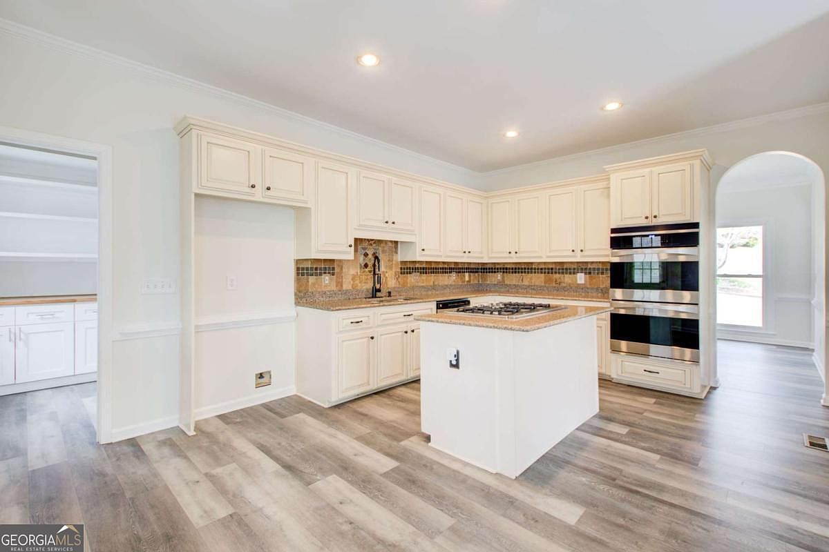 This bright and spacious kitchen features classic white cabinetry, granite countertops, and a central island equipped with a gas cooktop. The room is accented by a decorative tile backsplash and stainless steel double wall ovens, all set against modern wood-look flooring. The perspective is a wide-angle shot from the kitchen floor, showcasing the open layout and seamless flow into the adjacent rooms.
