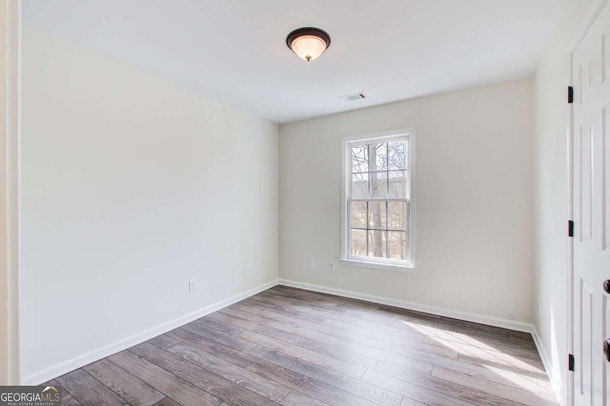 This is a bright, minimalist bedroom featuring light-colored walls and modern wood-look laminate flooring. A single window provides natural light and a view of the outdoors, while a simple ceiling light fixture illuminates the room. The space is clean and neutral, offering a versatile canvas for any interior design style.