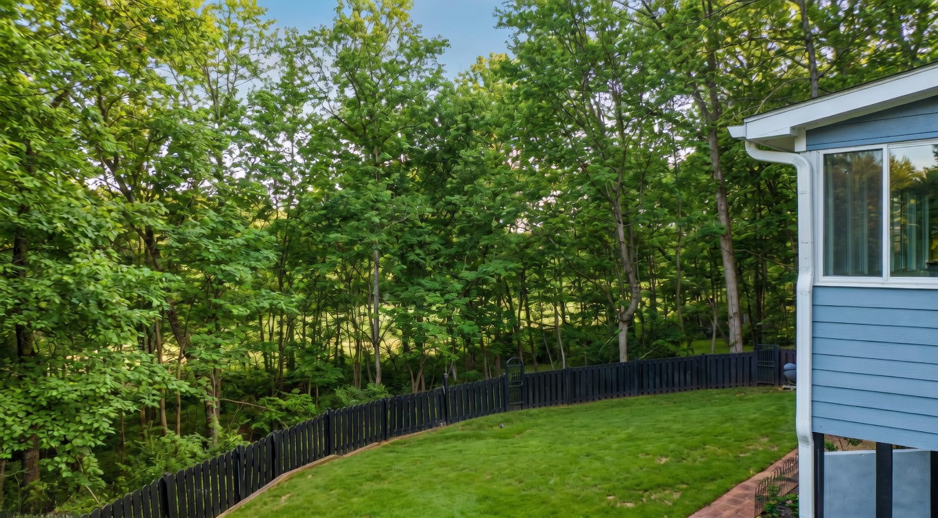 This elevated rear view captures a spacious backyard featuring a well-maintained lawn bordered by a dark wooden fence. Beyond the fence, a dense forest of tall, leafless trees creates a private, natural backdrop, suggesting a quiet and serene setting. The perspective from an upper level highlights the home's blue siding and large windows, emphasizing the connection between the indoor living space and the expansive outdoor environment.
