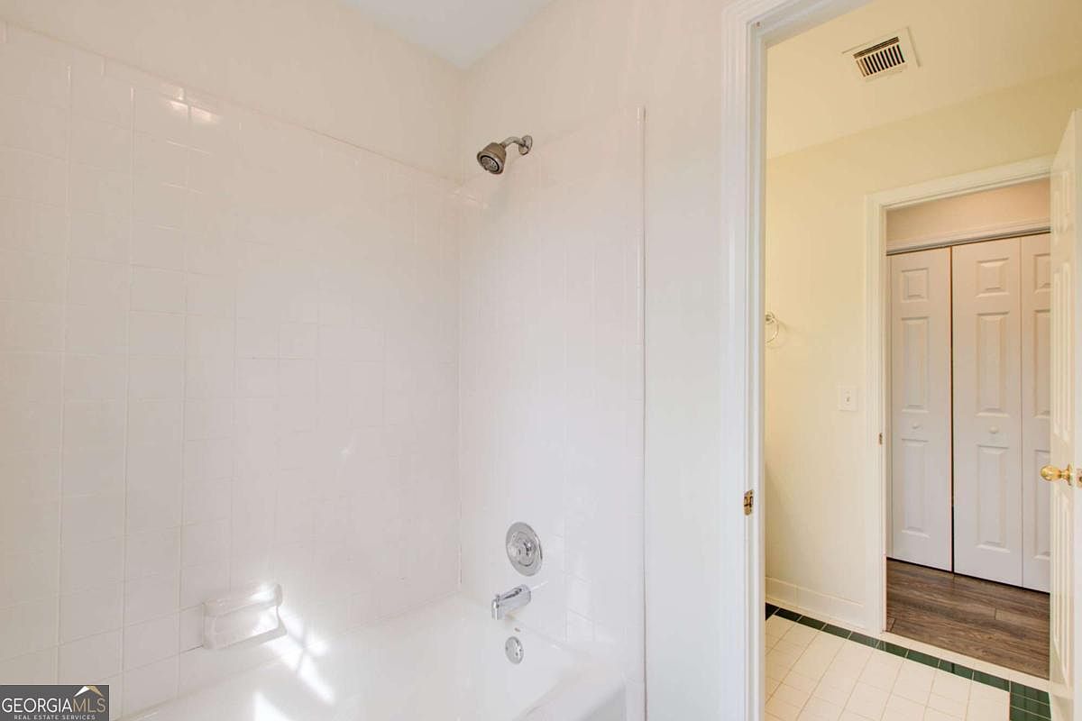 This image shows a clean, functional bathroom featuring a white tiled shower surround with a built-in soap dish and a standard showerhead. The perspective looks into the shower area from the adjacent hallway, which features white tiled flooring and a view of a closet with double doors. The space is bright and neutral, presenting a well-maintained and simple aesthetic suitable for a guest or secondary bathroom.