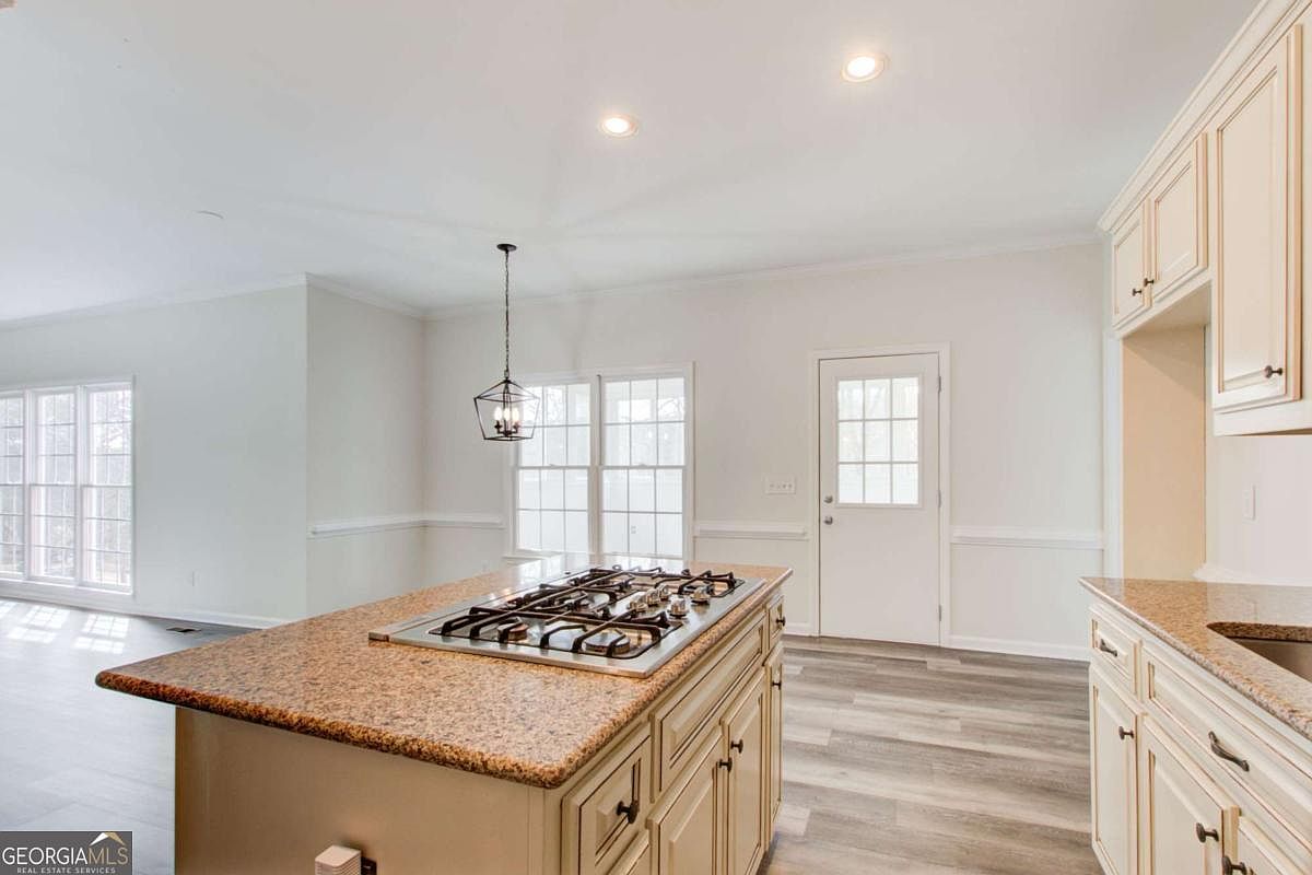 This bright and spacious kitchen features a large central island topped with speckled granite and a built-in gas cooktop. The room is accented by cream-colored cabinetry, light-toned wood-look flooring, and a classic pendant light hanging over the dining area. A glass-paned door and large windows provide ample natural light, creating an inviting and functional atmosphere.