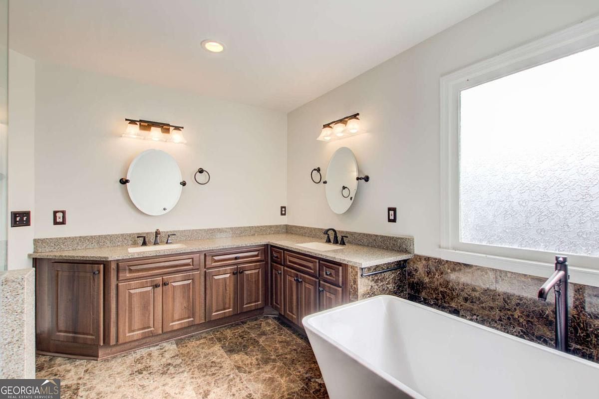 This spacious primary bathroom features a large L-shaped wooden vanity with dual sinks, granite countertops, and two oval mirrors. A deep, freestanding soaking tub is positioned near a large window, complemented by dark marble-style tile flooring and wall accents. The room is well-lit with modern vanity lighting, creating a clean and luxurious atmosphere.