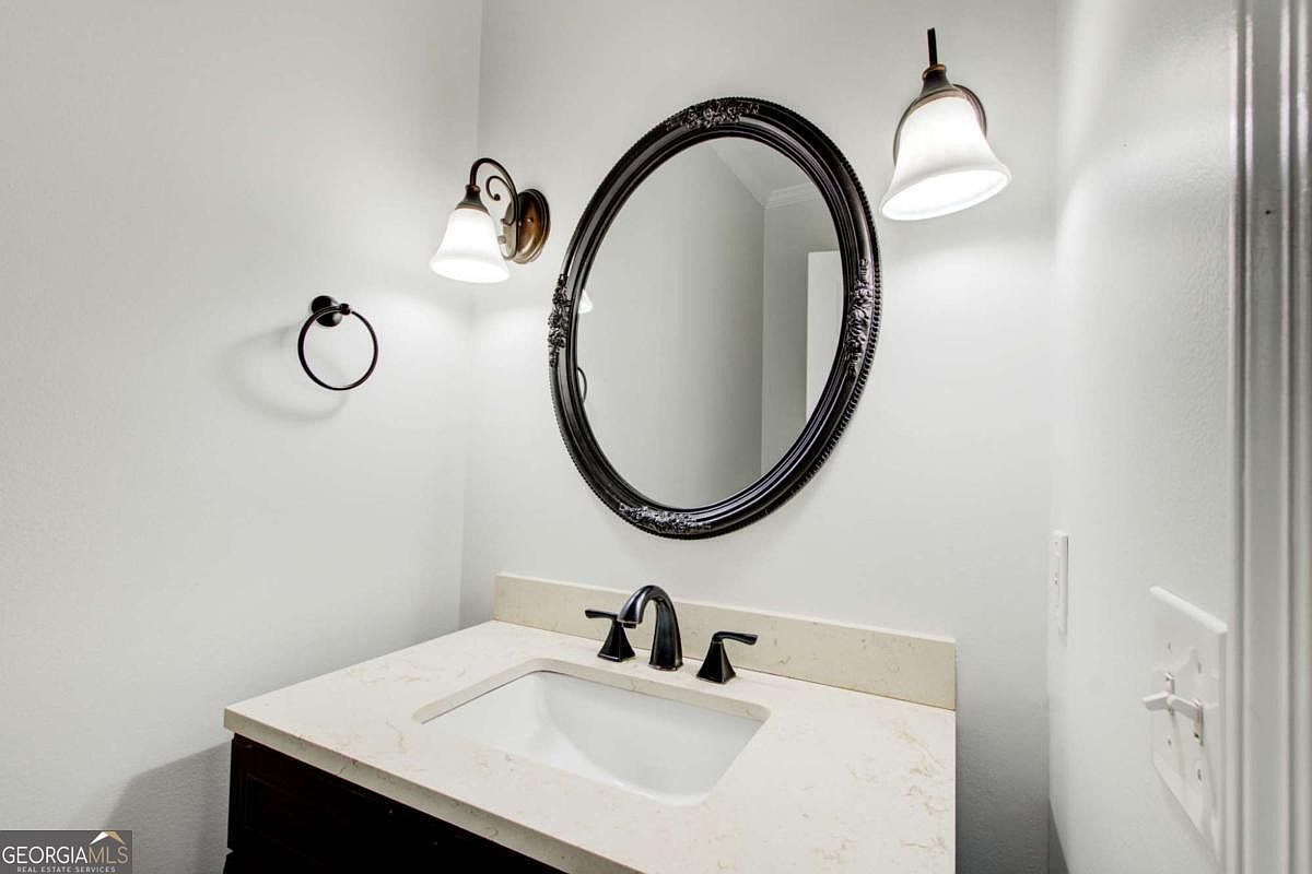 This is a clean and compact powder room featuring a dark wood vanity topped with a light-colored stone countertop and a modern dark-finished faucet. An ornate, oval-shaped black-framed mirror hangs above the sink, flanked by two elegant wall-mounted light fixtures. The space is finished with neutral walls and a simple towel ring, creating a polished and functional aesthetic.