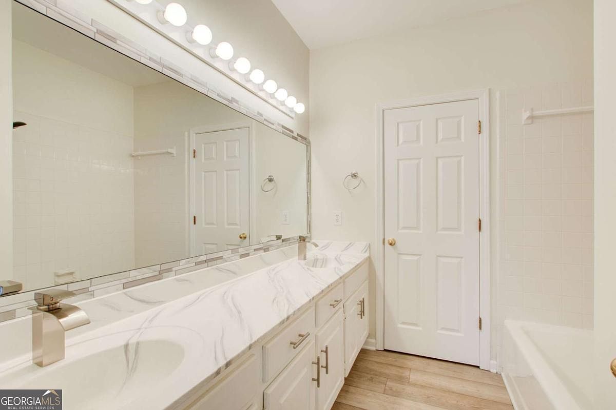 This bright and clean primary bathroom features a long vanity with a white marble-patterned countertop, dual sinks, and modern brushed-nickel fixtures. A large, expansive mirror spans the wall above the vanity, complemented by a multi-bulb vanity light fixture, while a white door leads to the rest of the home. The space is finished with light-toned wood-look flooring and neutral wall paint, creating a fresh and inviting atmosphere.