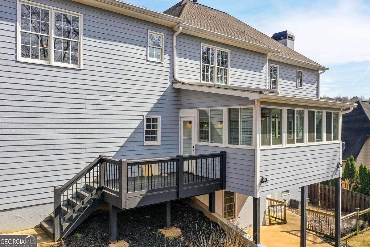 This rear view of a multi-story residential home showcases a light blue horizontal siding exterior with a prominent sunroom addition. A dark-painted wooden deck with stairs leads down to a lower-level concrete patio area, providing a functional outdoor living space. The perspective captures the architectural depth of the home, highlighting the transition between the elevated deck and the ground-level backyard.
