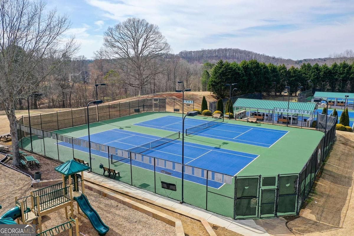 This high-angle aerial view showcases a well-maintained community tennis court facility featuring two blue-surfaced courts enclosed by chain-link fencing. Adjacent to the courts is a covered pavilion with seating, and a children's playground structure is visible in the foreground, all set against a backdrop of mature trees and rolling hills. The scene conveys a sense of active, family-friendly community living.