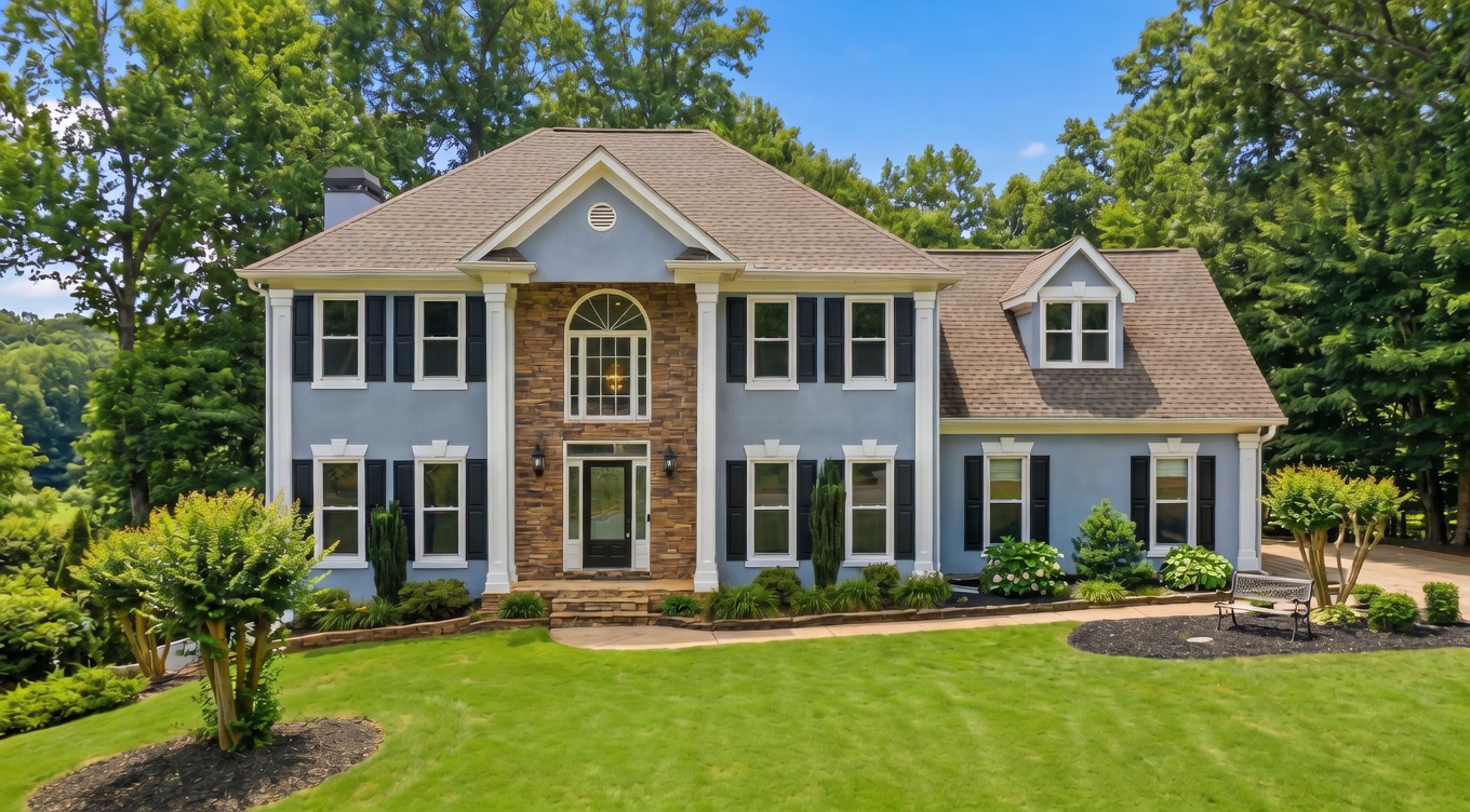 This elegant two-story craftsman-style home features a striking blue stucco exterior accented by a stone-faced central entryway with towering white columns. The property showcases meticulous landscaping, including mature trees and neatly defined garden beds, paired with a charming front lawn and a classic park bench. The inviting front facade, with its balanced architectural details and large windows, presents a high-end, welcoming appearance ideal for residential appeal.