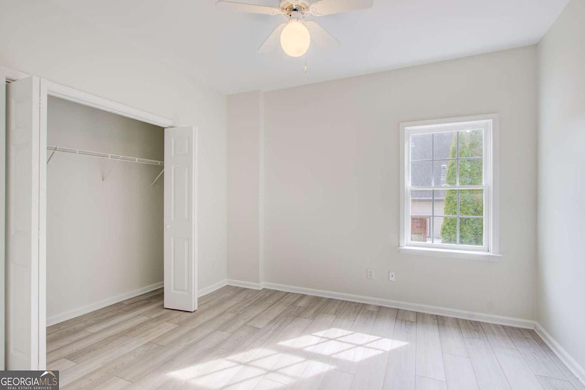 This bright and neutral bedroom features light-toned wood-look flooring and crisp white walls, creating a clean and inviting atmosphere. The room includes a ceiling fan with a central light fixture and a built-in closet with double doors. A single window allows natural light to filter in, offering a view of the exterior surroundings.