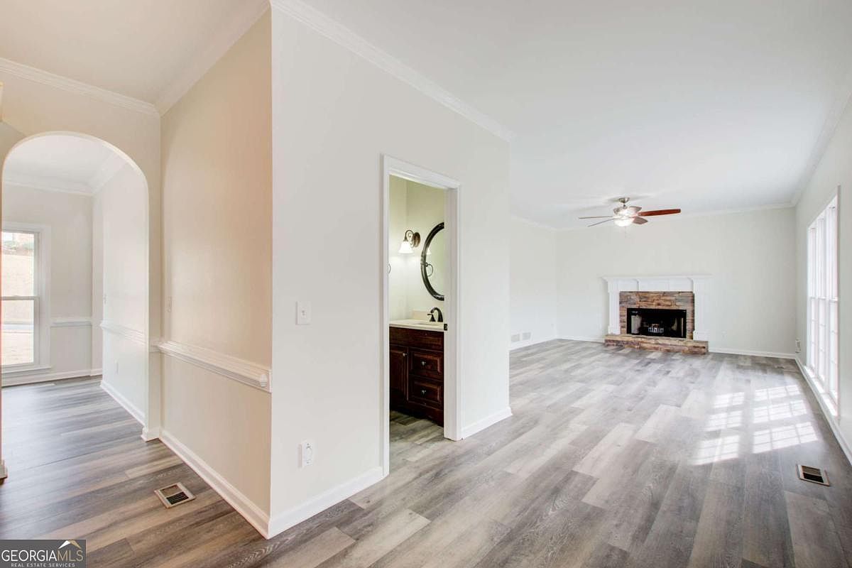 This bright and open living area features modern wood-look flooring and neutral walls, creating a clean and inviting atmosphere. A stone-faced fireplace serves as a focal point, while an adjacent doorway provides a glimpse into a small bathroom. The perspective is from a hallway looking into the main living space, emphasizing the home's open floor plan and natural light.