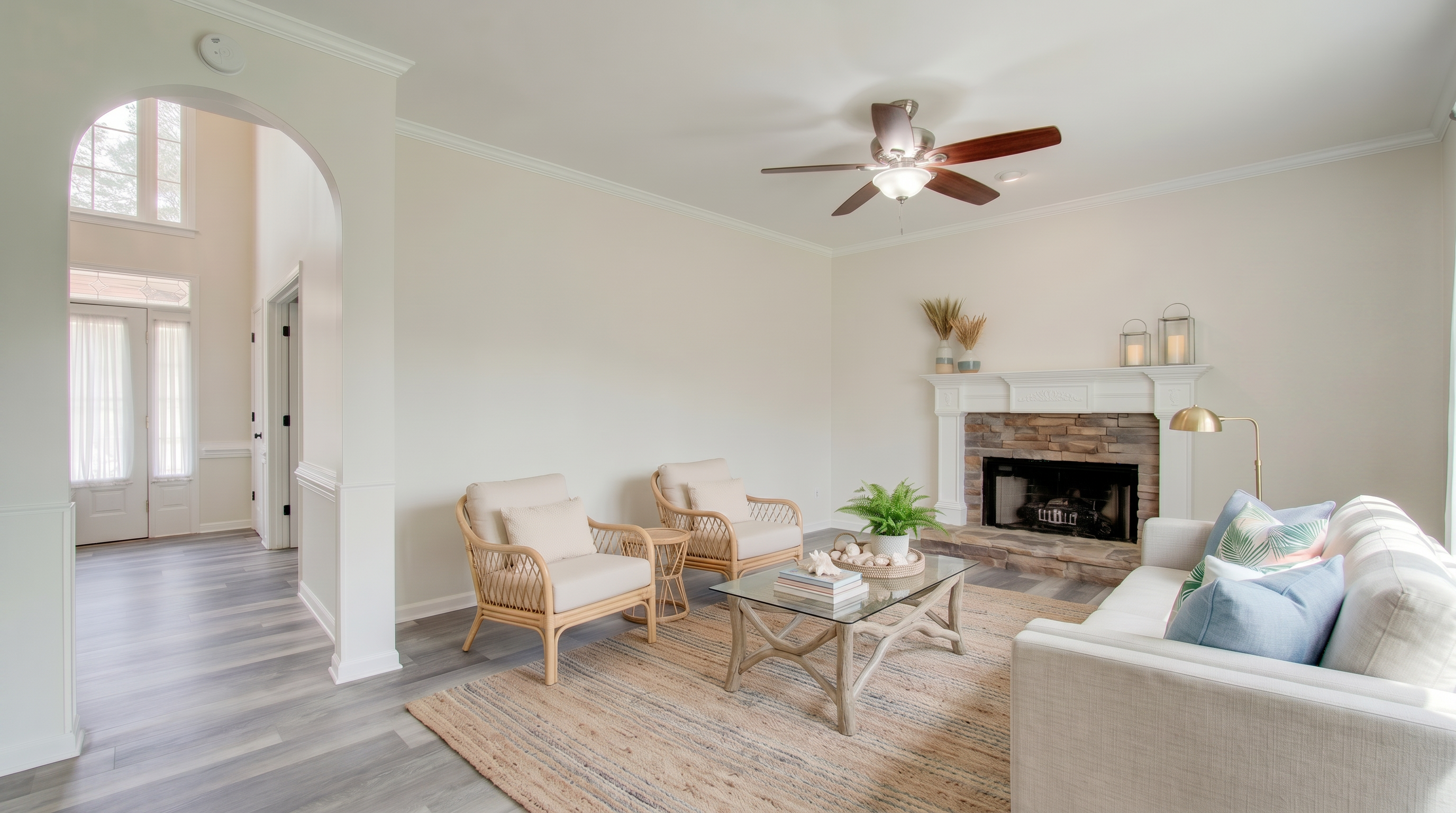 This bright and inviting living room features a stone fireplace as a central focal point, complemented by light-toned neutral furniture and a natural area rug. The space includes two rattan-style accent chairs and a comfortable sofa, creating a cozy and airy atmosphere. A large, well-lit entryway with arched architectural details provides a seamless transition to the rest of the home, emphasizing an open-concept layout.