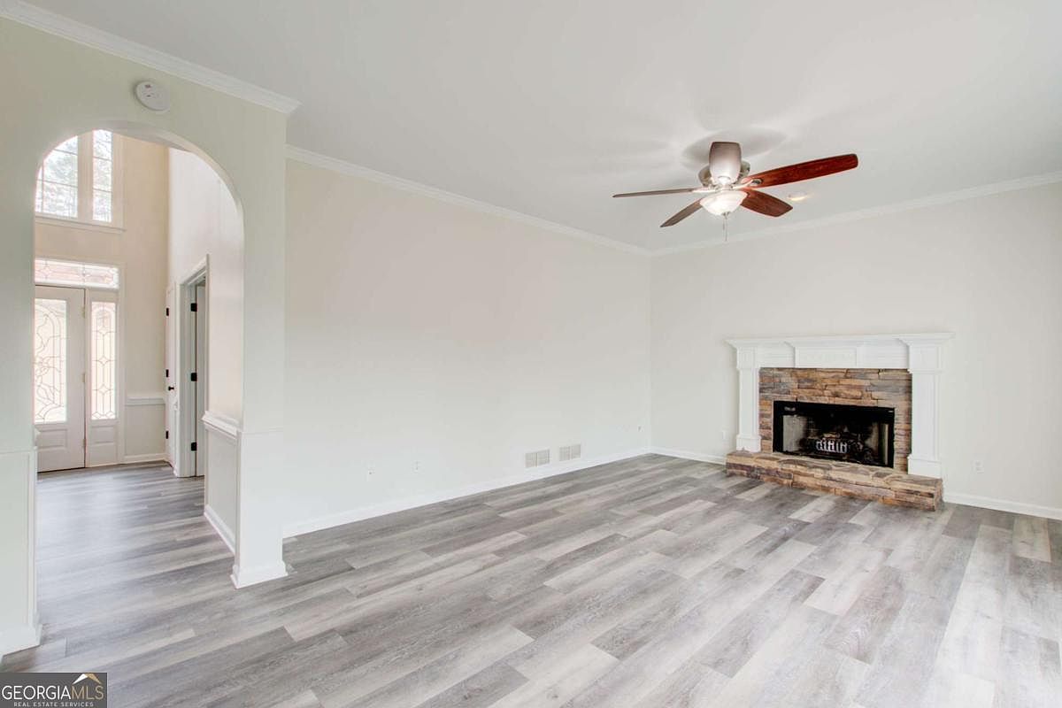 This spacious living room features light-colored walls and modern wood-look flooring, creating a bright and airy atmosphere. A prominent stone-faced fireplace with a white mantel serves as the focal point, while an arched entryway leads to a high-ceilinged foyer. The room is illuminated by a ceiling fan with integrated lighting, offering a clean and neutral canvas for potential buyers.
