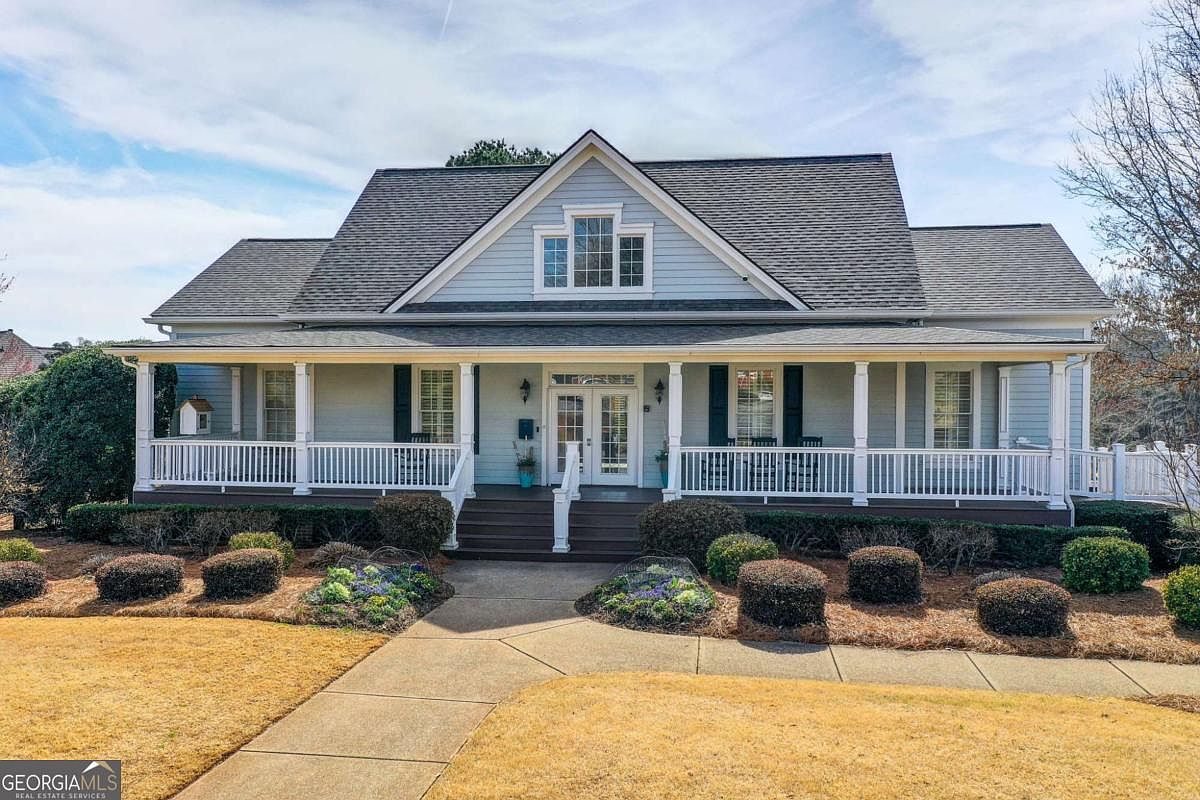 This charming, light blue craftsman-style home features a welcoming full-width front porch with white railings and columns. The symmetrical facade includes a central gable with a triple-window arrangement, dark shutters, and a concrete walkway leading to the double-door entryway. The landscaping is neatly manicured with rounded shrubs and flower beds, creating a warm and inviting curb appeal.