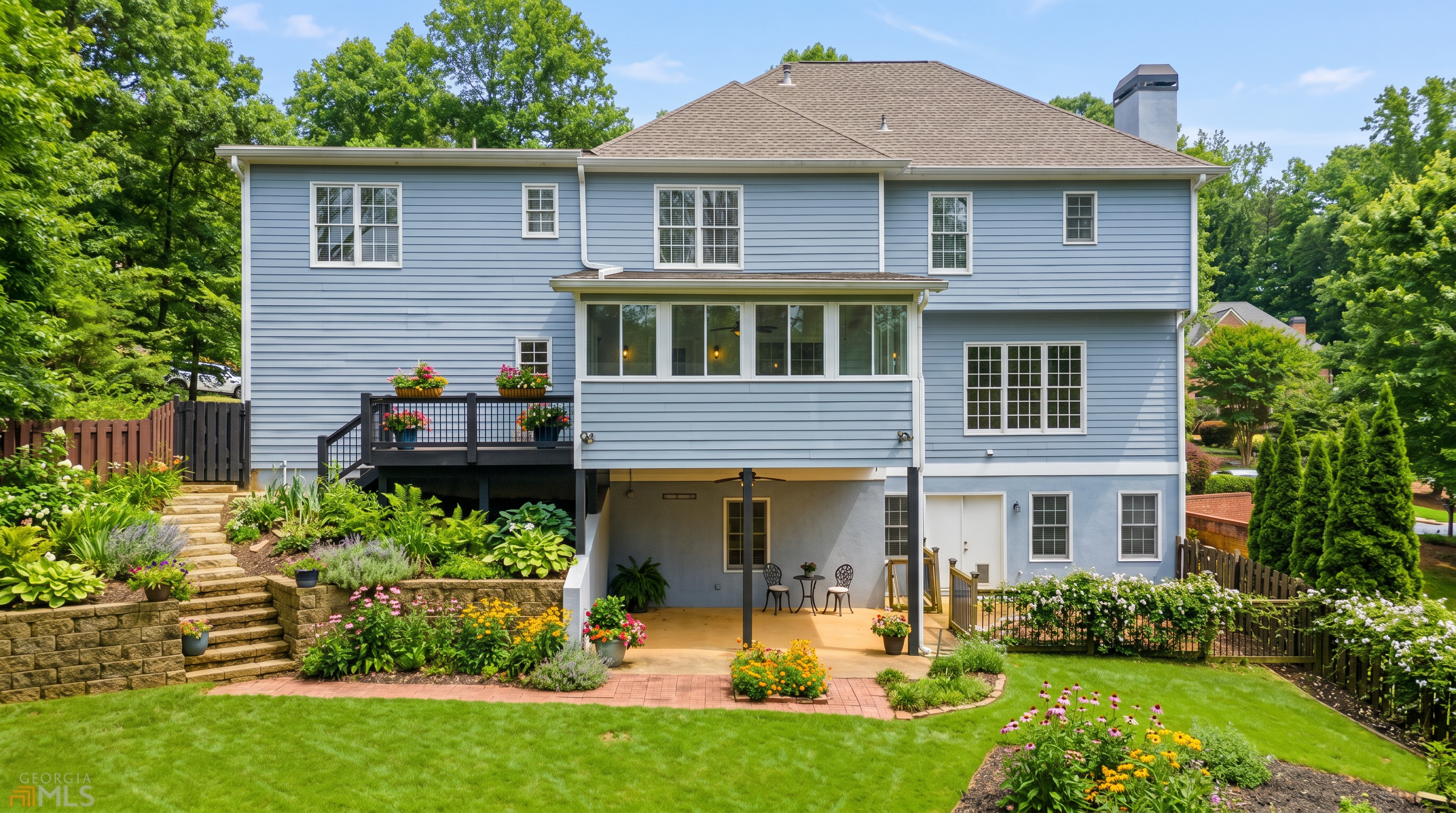 This rear view of a multi-story suburban home showcases a light blue siding exterior with a prominent elevated deck and a covered patio area below. The backyard features a tiered stone retaining wall, a brick-paved walkway, and a fenced-in lawn, creating a well-defined outdoor living space. The perspective is captured from a slightly elevated outdoor vantage point, highlighting the home's architectural layout and its integration with the landscaped grounds.