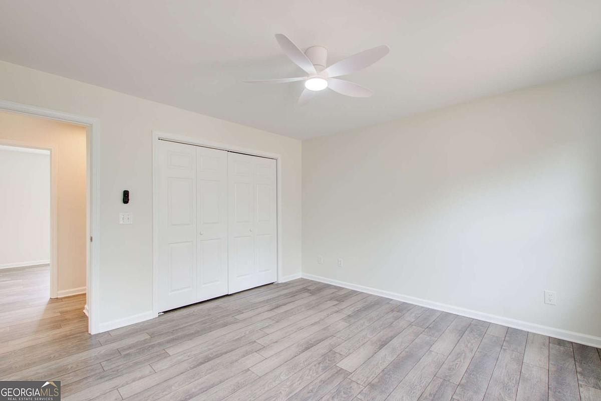 This bright and neutral bedroom features light-colored walls and modern wood-look laminate flooring. A white ceiling fan with integrated lighting is centered in the room, while a set of white bi-fold closet doors provides ample storage. The perspective is from the doorway, looking into the clean, minimalist space that offers a versatile canvas for any decor style.