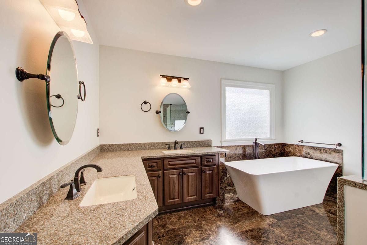 This spacious primary bathroom features a double vanity with dark wood cabinetry, granite countertops, and two oval mirrors. A large, freestanding white soaking tub sits prominently in the corner, complemented by dark marble-style flooring and a frosted window that allows for natural light while maintaining privacy. The room offers a luxurious and clean aesthetic, perfect for a spa-like retreat.