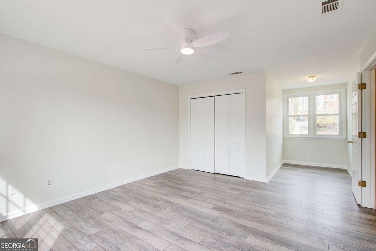 This bright and neutral bedroom features light-colored walls and modern wood-look laminate flooring. The room includes a ceiling fan, a double-door closet, and a window that allows for natural light, creating a clean and versatile living space. The perspective is from the doorway, looking into the room to showcase its layout and size.