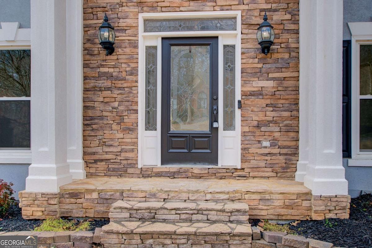 This inviting front entryway features a dark-framed glass door flanked by sidelights and topped with a transom window, all set against a textured stone veneer facade. Two classic black lanterns provide elegant lighting, while the stone steps lead up to a welcoming porch area. The overall aesthetic is sophisticated and traditional, creating a strong sense of curb appeal.