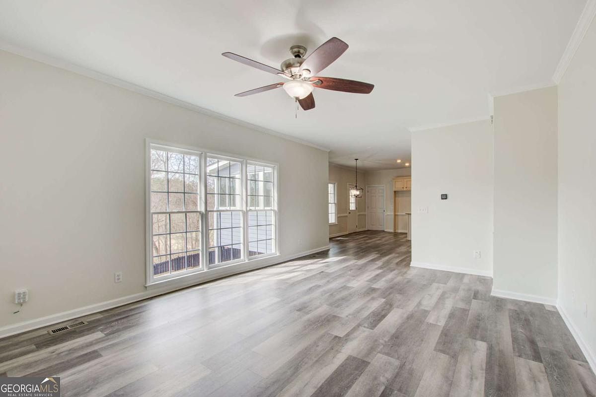 This spacious, open-concept living area features light-toned walls and modern wood-look luxury vinyl plank flooring. A large window assembly allows for ample natural light, while a ceiling fan with a light fixture serves as the central focal point. The room flows seamlessly into an adjacent dining or kitchen area, creating an airy and inviting atmosphere suitable for versatile furniture arrangements.