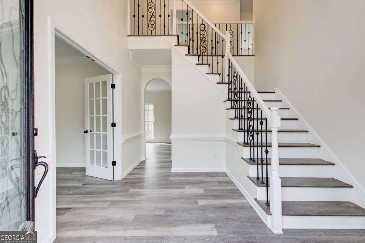 This bright and airy foyer features a grand staircase with dark wood treads and white risers, complemented by elegant black wrought-iron balusters. The space is finished with light-toned wood-look flooring and crisp white walls, creating a clean and inviting entrance. A French door leads to an adjacent room, while an arched doorway provides a view into the deeper parts of the home, emphasizing an open and flowing floor plan.