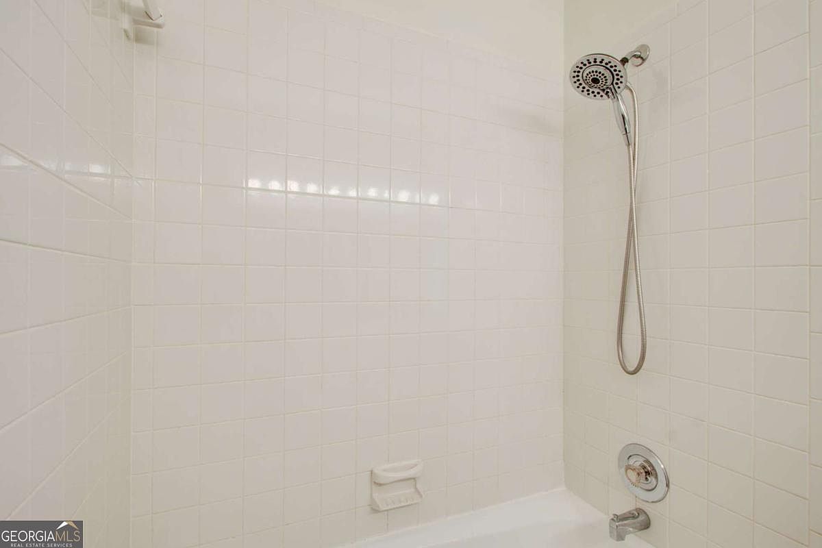 This image shows a standard bathtub and shower combination featuring white square wall tiles and a chrome showerhead fixture. The space is clean and functional, with a built-in soap dish integrated into the tiled wall. The perspective is a straight-on view of the shower enclosure, highlighting the neutral and simple design.