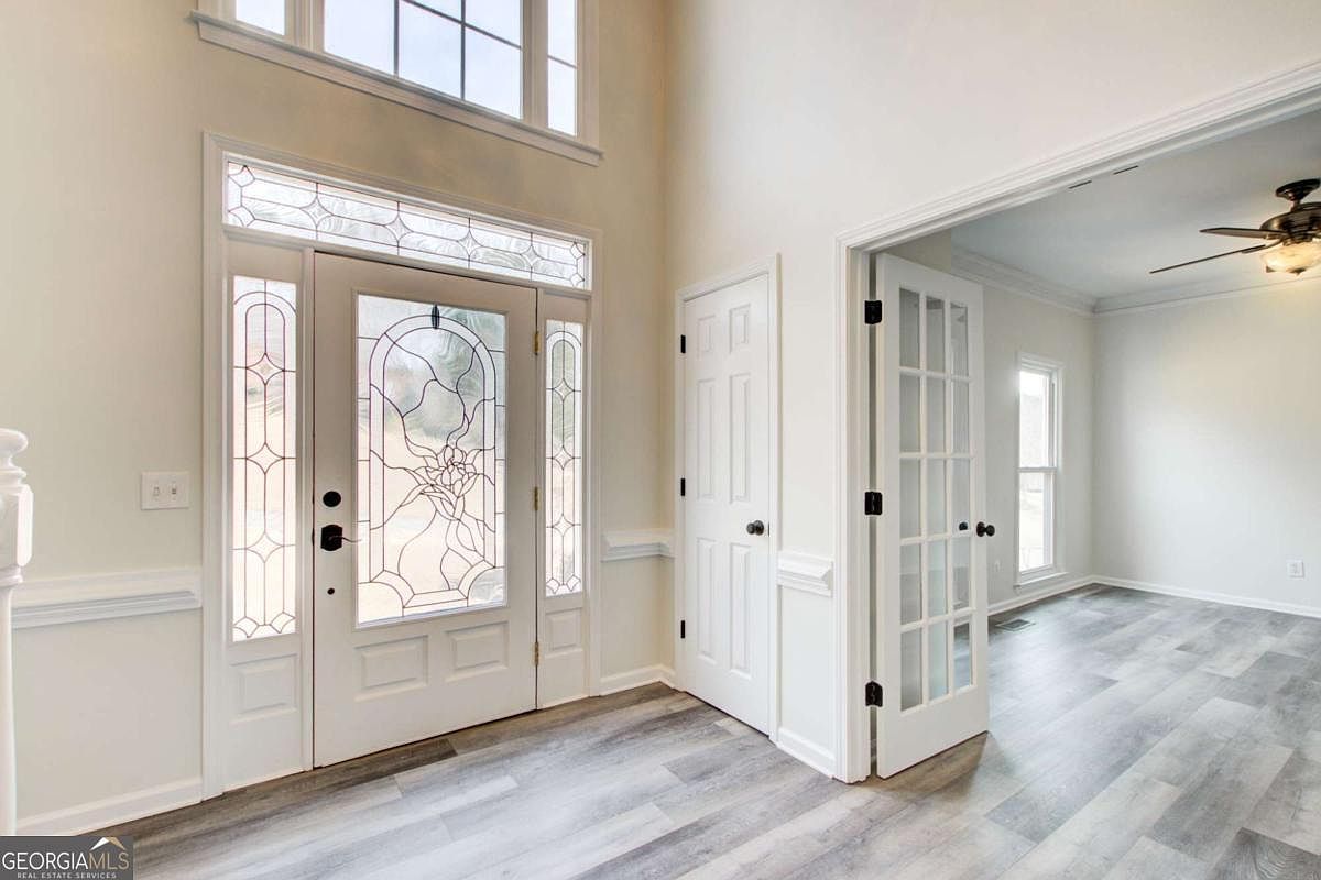 This bright and airy entryway features a grand two-story ceiling with a large transom window above a decorative glass-paneled front door. To the right, French doors open into an adjacent room, while light-toned wood-look flooring flows throughout the space. The clean white walls and classic trim create a welcoming and sophisticated first impression for potential buyers.