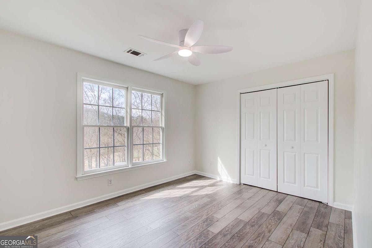 This bright and neutral bedroom features light-colored walls and modern wood-look flooring, creating a clean and inviting atmosphere. The room is equipped with a white ceiling fan and a double-door closet, while a large window allows for ample natural light and a view of the outdoors. The perspective is a wide-angle shot from the corner of the room, showcasing the simple, versatile layout.