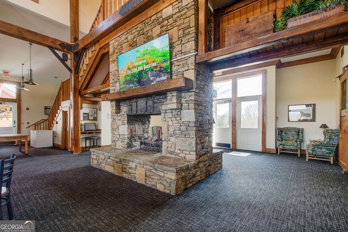 This spacious, rustic-style clubhouse interior features a prominent floor-to-ceiling stone fireplace as its central focal point, topped with a large television. The room is characterized by exposed timber beams, high ceilings, and a neutral color palette, creating a warm and inviting atmosphere. The space includes comfortable seating areas, a staircase leading to an upper level, and large windows that allow for ample natural light, making it an ideal communal gathering area.