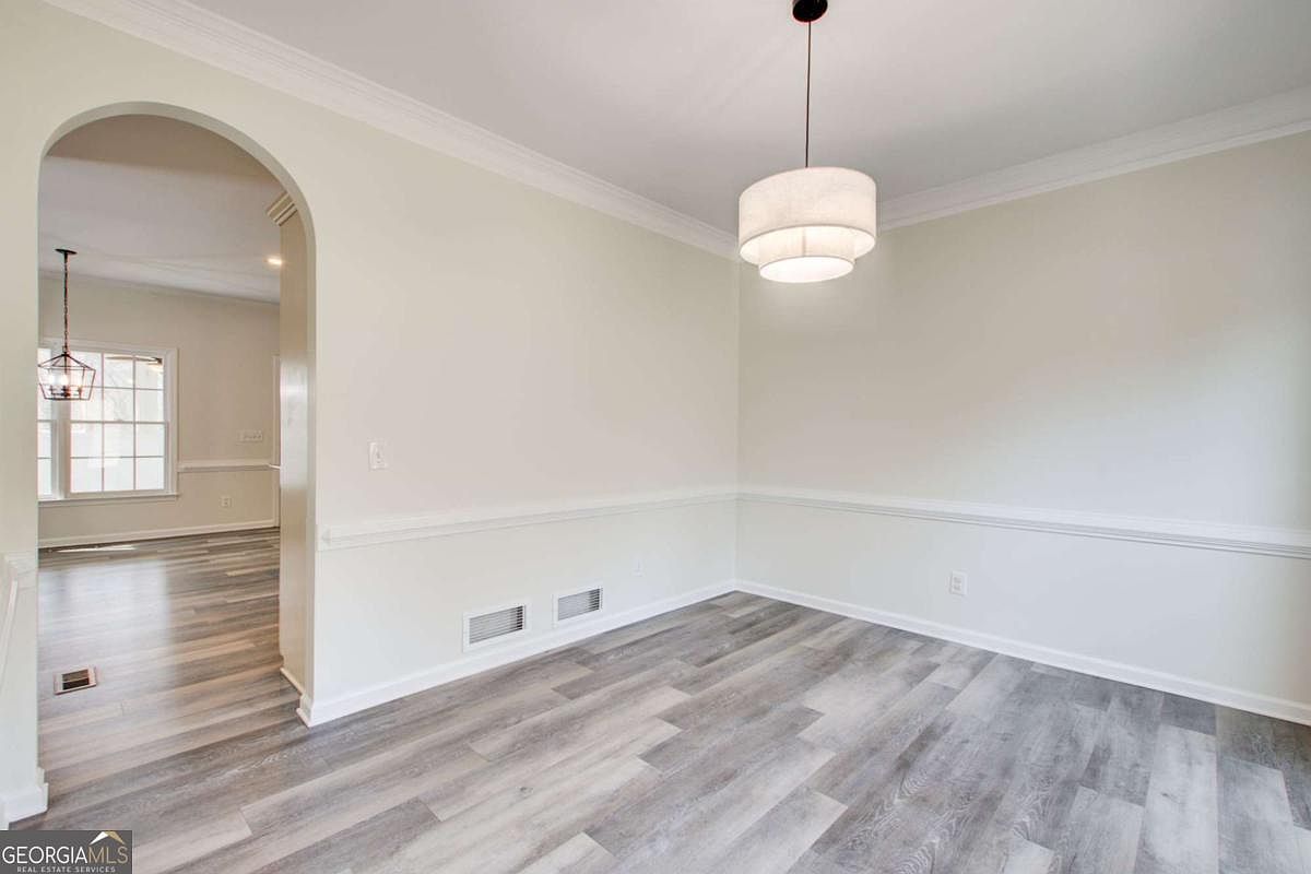 This bright and airy dining room features modern light-gray wood-look flooring and neutral walls accented by classic chair rail molding. A contemporary tiered drum pendant light serves as the focal point, while an arched entryway provides a seamless transition into the adjacent living space. The room offers a clean, minimalist aesthetic that is ready for personalization.