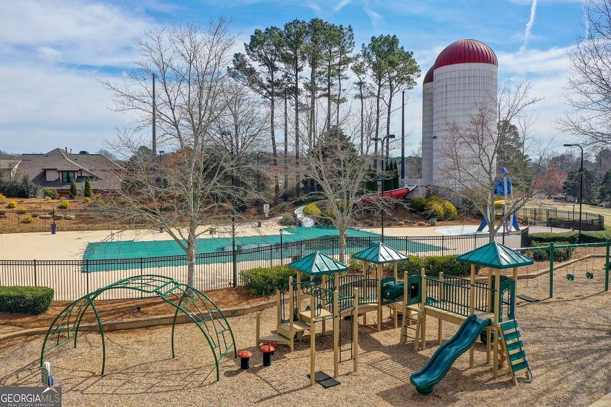 This elevated view captures a vibrant community amenity area featuring a playground with climbing structures and slides, situated adjacent to a fenced-in swimming pool covered for the season. In the background, two prominent silo-style structures stand out against a clear blue sky, surrounded by mature trees and a residential building. The scene conveys a family-friendly atmosphere, highlighting the recreational facilities available to residents.