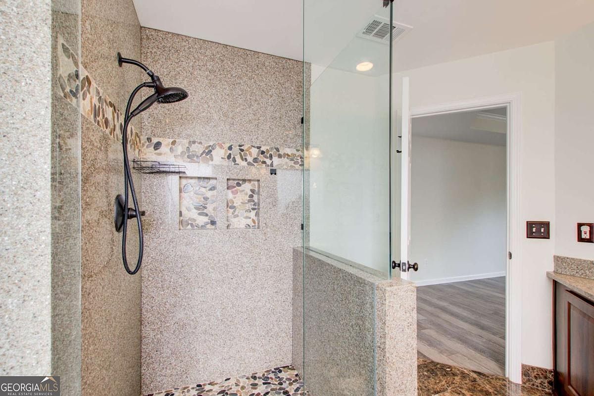 This primary bathroom features a spacious walk-in shower enclosed by a frameless glass panel, showcasing a textured granite-style wall finish and a decorative pebble-tile accent strip. The shower includes a modern dark-finished showerhead and built-in recessed shelving for storage. The perspective looks into the shower area, revealing a glimpse of an adjacent room with wood-look flooring through the open doorway.