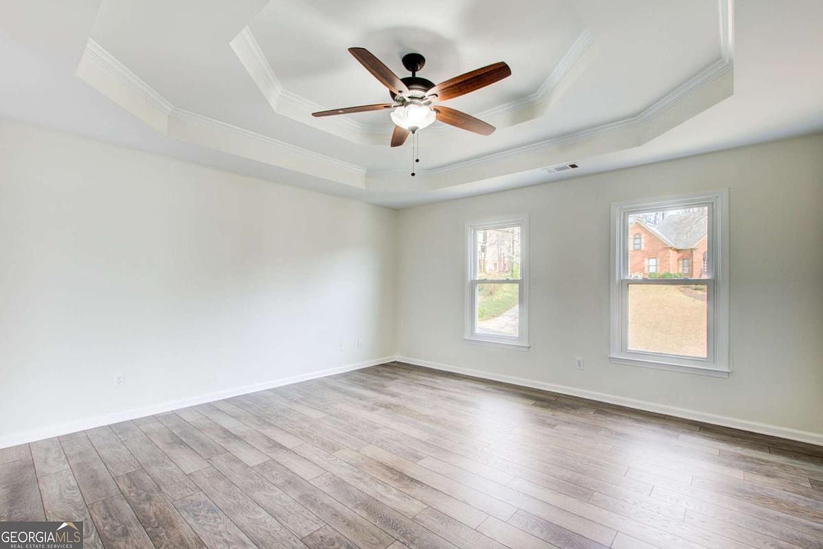 This spacious, unfurnished bedroom features light-colored walls and modern wood-look laminate flooring, creating a bright and airy atmosphere. A prominent tray ceiling with a central ceiling fan adds architectural interest, while two large windows provide natural light and a view of the exterior. The room offers a clean, neutral canvas ready for personalization.