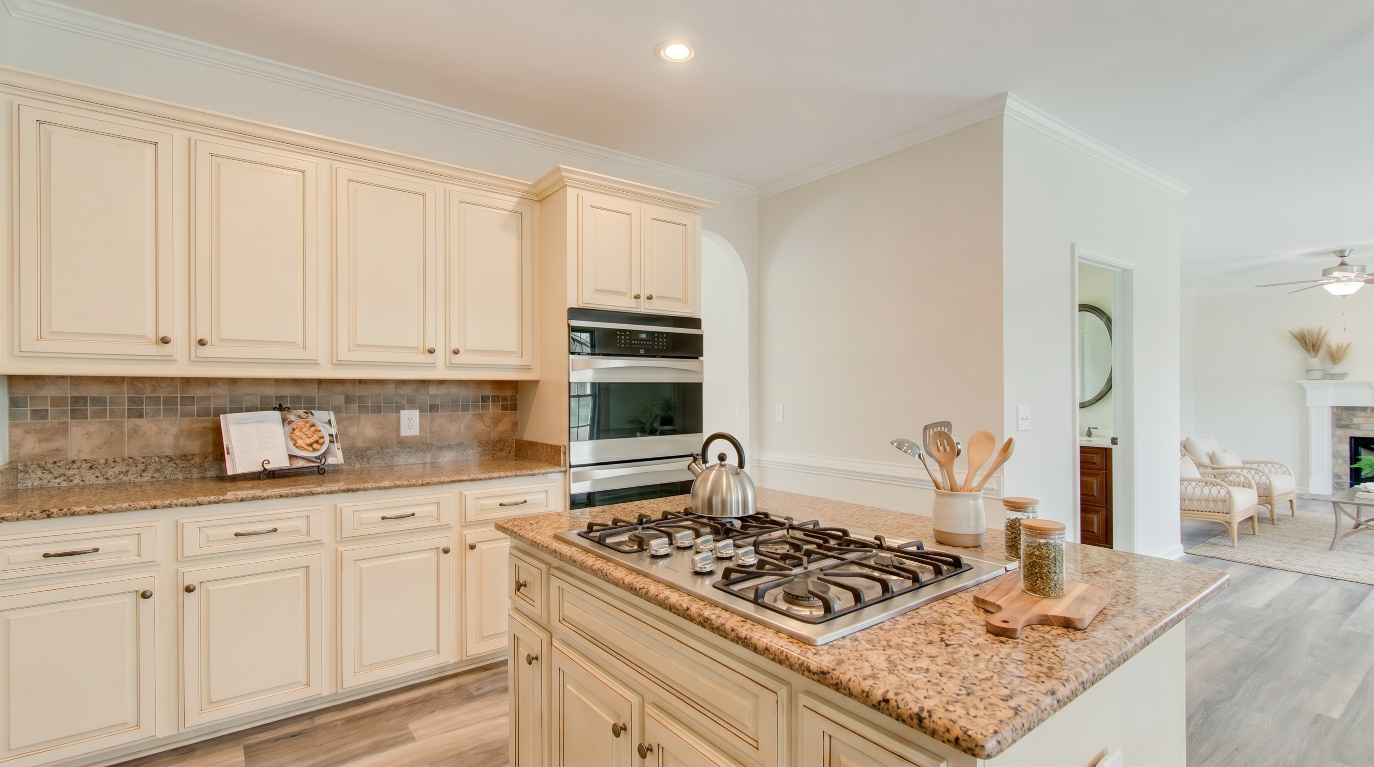 This spacious kitchen features off-white cabinetry, granite countertops, and a central island equipped with a gas cooktop. The warm color palette creates a welcoming atmosphere, complemented by high-end stainless steel wall ovens and modern wood-look laminate flooring that seamlessly transitions into the adjacent living space.