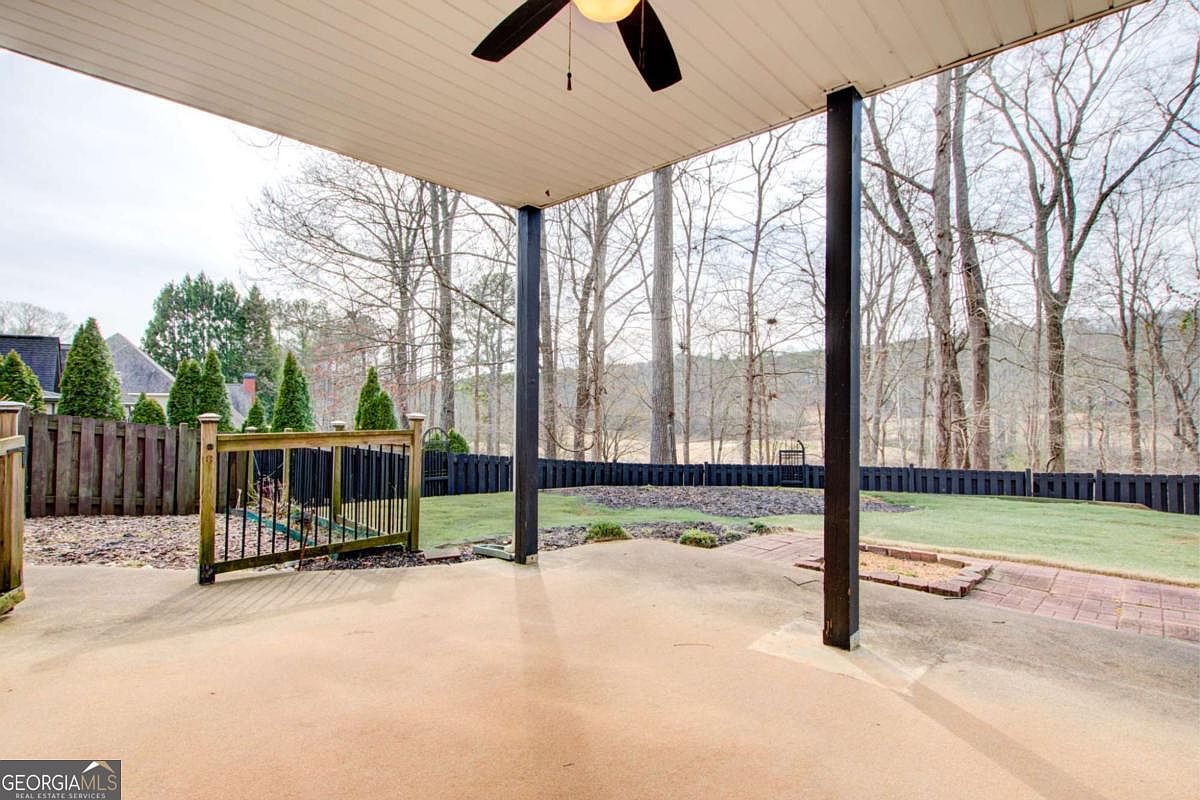 This image captures a covered concrete patio looking out over a private, fenced-in backyard. The space features a ceiling fan and provides a clear view of the lawn, mature trees, and a wooden privacy fence. The perspective is from under the patio roof, emphasizing the transition between the indoor-outdoor living area and the natural landscape beyond.