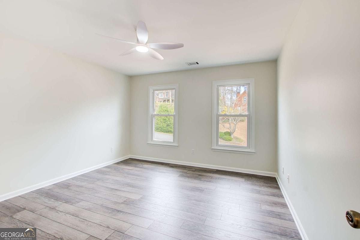 This bright and neutral bedroom features light-colored walls and modern wood-look laminate flooring. Two windows provide natural light, while a ceiling fan with integrated lighting serves as the central fixture. The room offers a clean, blank canvas ready for personalization.