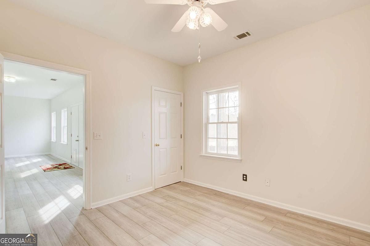 This bright and neutral bedroom features light-toned wood-look flooring and crisp white walls, creating a clean and versatile living space. A white door leads to a closet, while a window provides natural light, and a ceiling fan with a light fixture is centered above. The perspective looks into the room from a doorway, revealing a glimpse of an adjacent hallway with similar flooring.