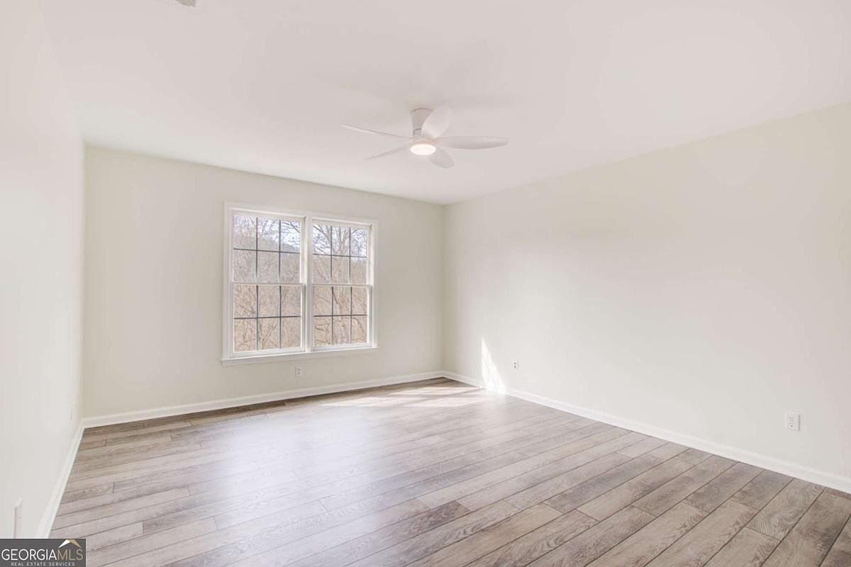 This is a bright, neutral-toned bedroom featuring light-colored walls and modern wood-look laminate flooring. The room is illuminated by a central ceiling fan with integrated lighting and natural light streaming through a large double window. The space is clean, unfurnished, and offers a versatile canvas for any interior design style.