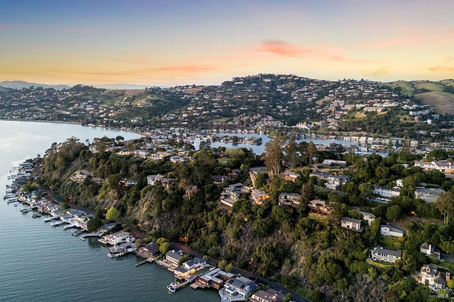 This high-angle aerial view captures a picturesque coastal hillside community at sunset, characterized by homes nestled among lush trees and steep terrain. The scene features a calm bay with numerous private docks and waterfront residences, transitioning into a dense residential area that climbs the rolling hills in the background. The warm, golden light of the setting sun creates a serene and inviting atmosphere, highlighting the unique topography and waterfront lifestyle of the location.