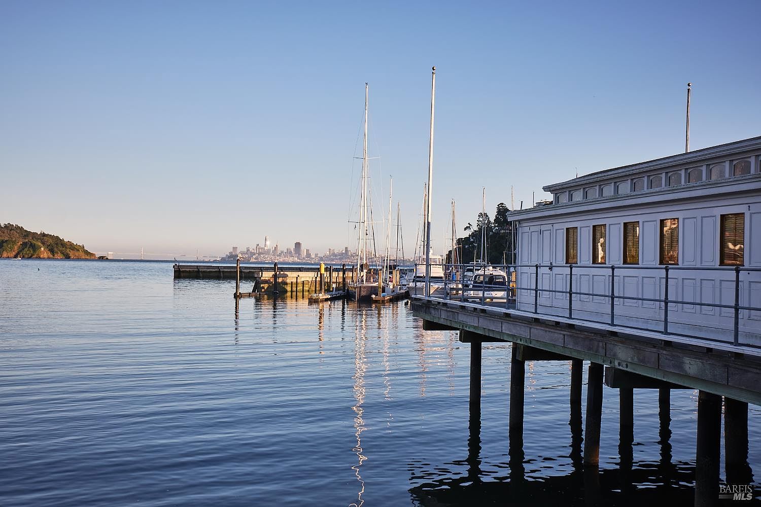 This scenic waterfront view captures a wooden pier extending over calm, reflective water, featuring a white, classic-style building with multiple windows. In the distance, the San Francisco skyline is visible under a clear blue sky, creating a serene and picturesque atmosphere. The perspective is from the water's edge, emphasizing the tranquil connection between the structure and the bay.