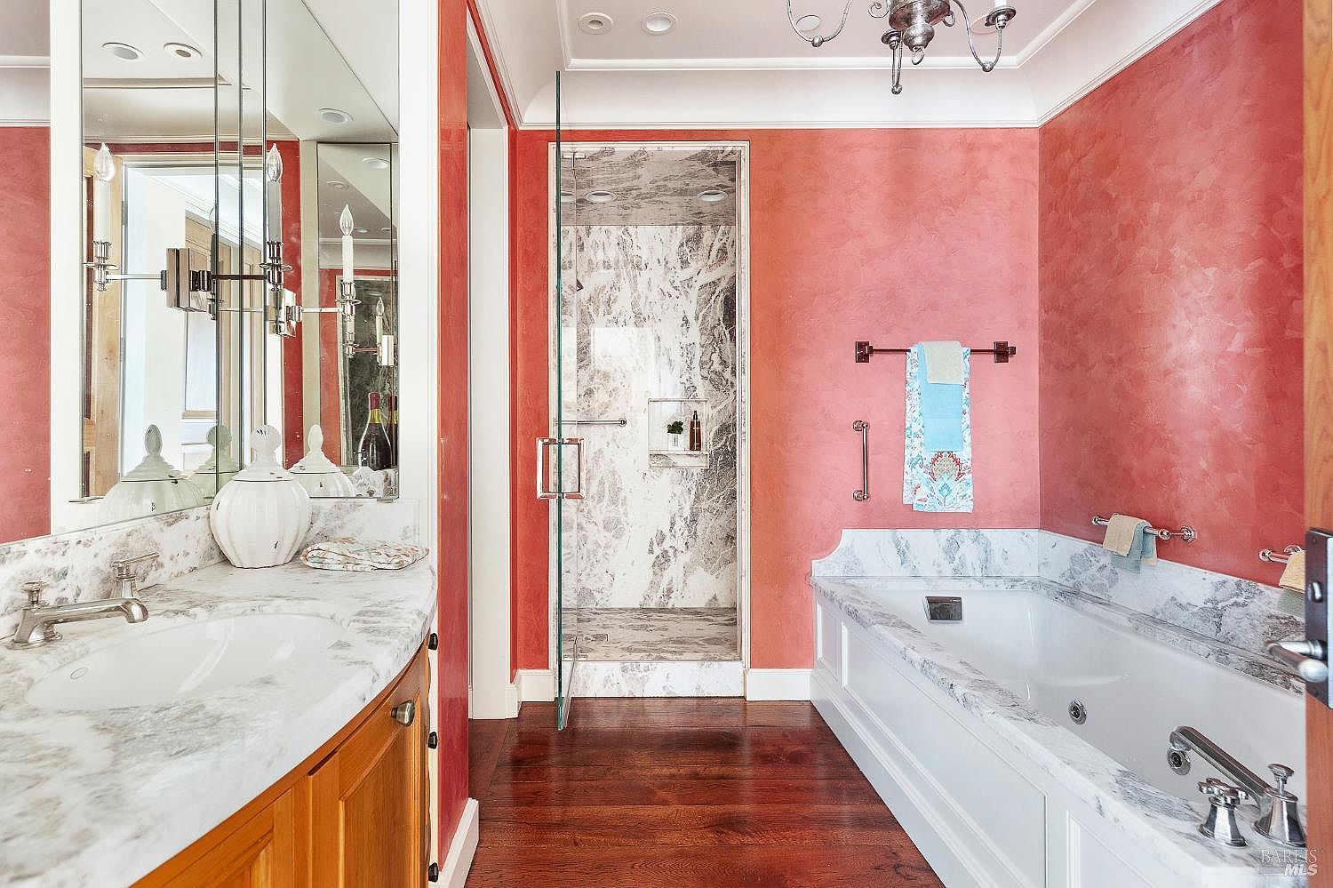 This primary bathroom features a warm, textured coral-toned wall finish paired with rich, dark wood flooring. The space includes a built-in soaking tub with a stone surround, a separate glass-enclosed shower with patterned stone walls, and a vanity with a stone countertop. A decorative chandelier hangs from the ceiling, adding a touch of elegance to the traditional design.