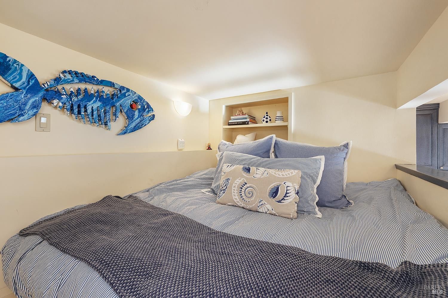 This cozy loft-style bedroom features a low-profile bed dressed in blue and white striped linens with a textured throw blanket. A decorative blue fish sculpture adorns the wall, while a small built-in recessed shelf provides storage for books and decor. The space is intimate and bright, utilizing a neutral color palette to create a relaxing, coastal-inspired atmosphere.