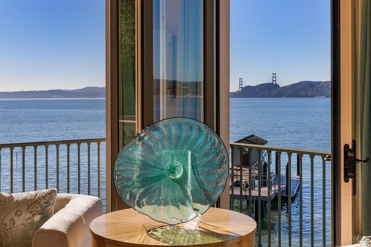 This interior view captures a serene living space featuring a round light wood table topped with a decorative teal glass art piece. Large windows provide an expansive, unobstructed view of the bay and the Golden Gate Bridge in the distance. The perspective looks out from a comfortable seating area toward a private balcony and the water, emphasizing the home's prime waterfront location.