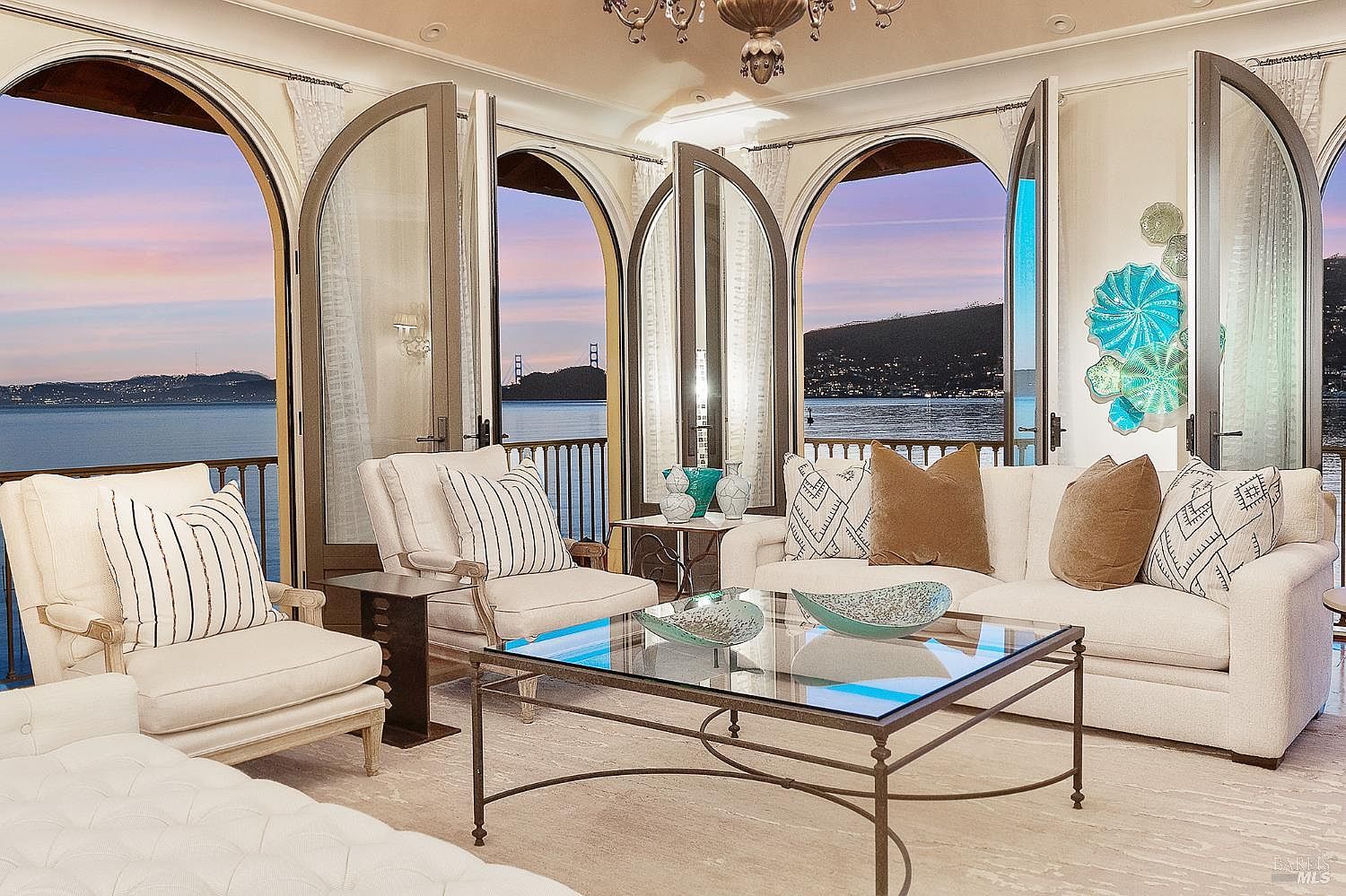 This elegant living room features a sophisticated neutral color palette with plush white seating, striped accent pillows, and a glass-topped coffee table. The space is defined by large arched glass doors that open onto a balcony, framing a stunning panoramic view of the bay and the Golden Gate Bridge at sunset. Decorative blue glass art pieces and a classic chandelier add a touch of refined luxury to the bright, airy atmosphere.