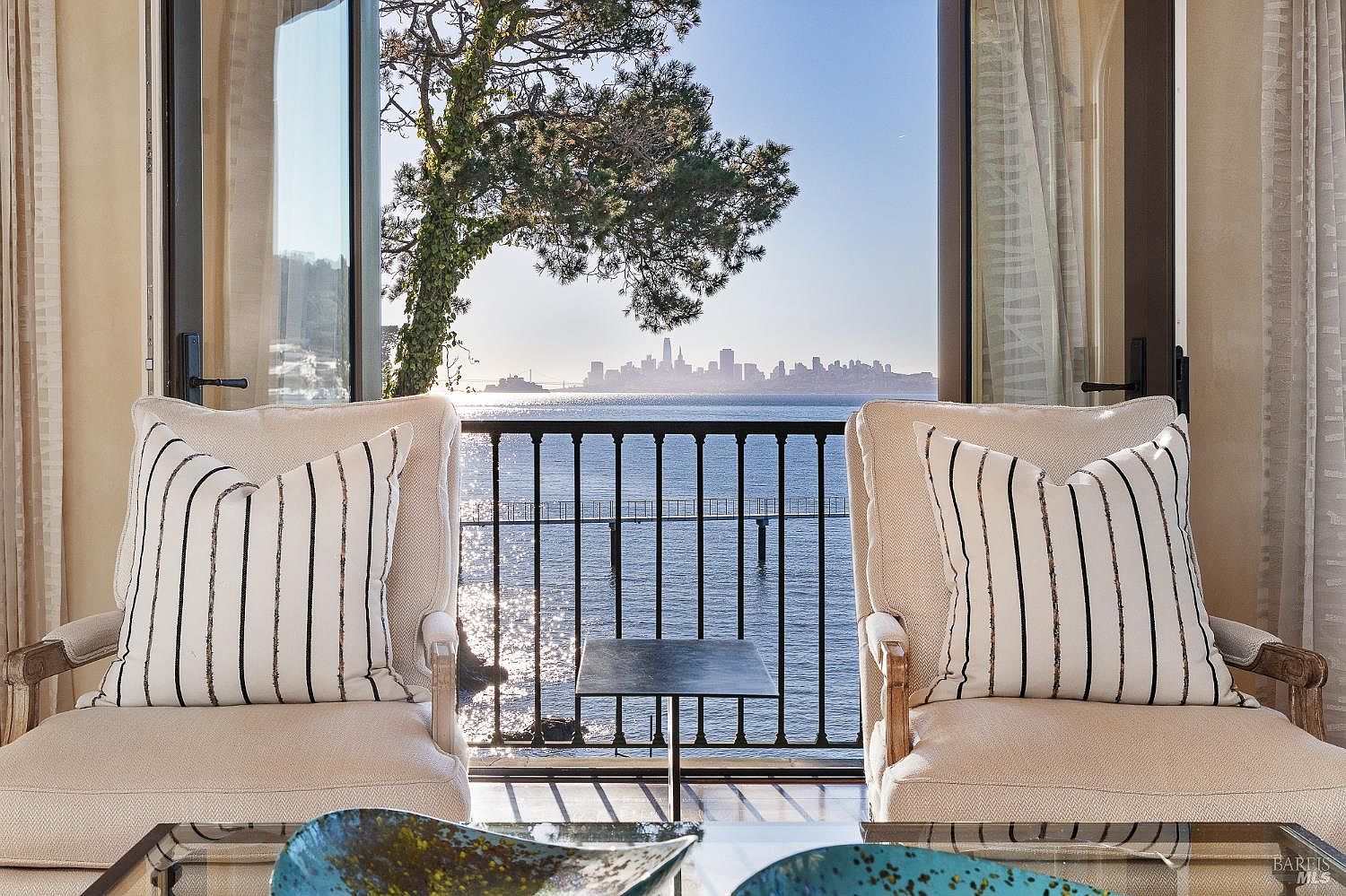 This interior space features two plush, light-colored armchairs with striped accent pillows, positioned to face a large opening that leads to a balcony. The view through the opening showcases a serene body of water with a distant city skyline and a prominent tree framing the scene. A small, dark metal side table sits between the chairs, and a decorative bowl is visible in the foreground, creating a sophisticated and tranquil atmosphere.