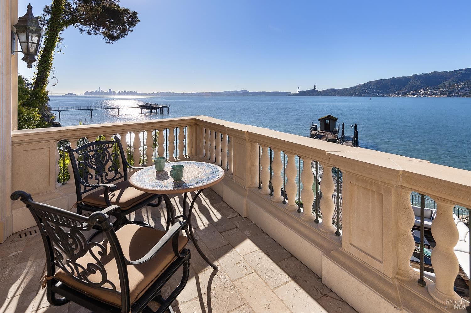 This elevated balcony offers a stunning, panoramic view of the bay, featuring a classic stone balustrade and light-colored stone flooring. A small, round mosaic-topped table with two ornate metal chairs provides an intimate seating area, perfect for enjoying the waterfront scenery. The perspective captures the serene atmosphere, highlighting the expansive water view, a distant city skyline, and a private dock below.