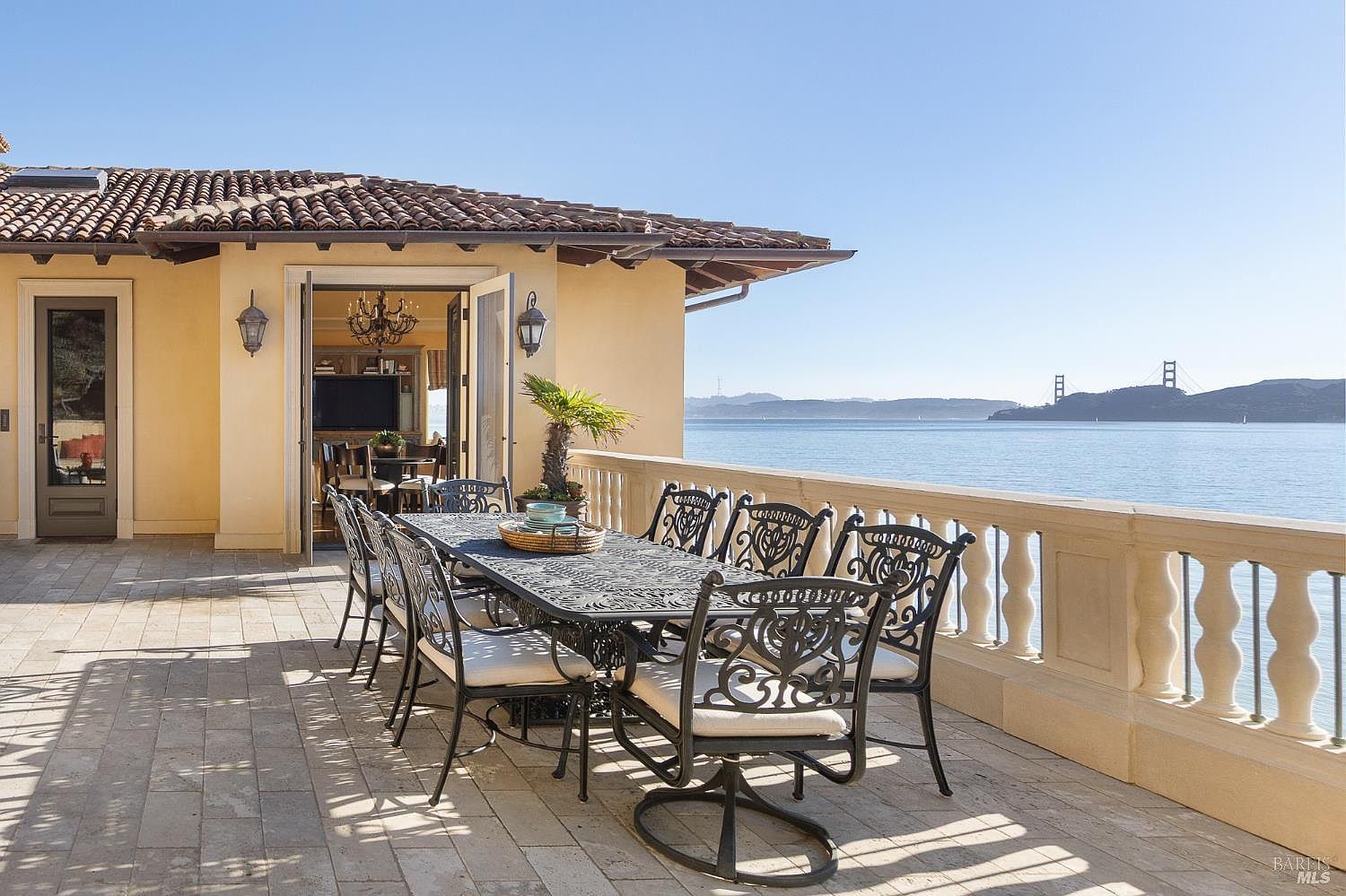 This expansive outdoor terrace features a large, ornate metal dining set positioned to overlook a stunning waterfront view, including the Golden Gate Bridge in the distance. The space is paved with light-toned stone tiles and bordered by a classic balustrade, creating a sophisticated setting for al fresco dining. Large glass doors open from the terrace into the home's interior, seamlessly blending indoor and outdoor living areas.