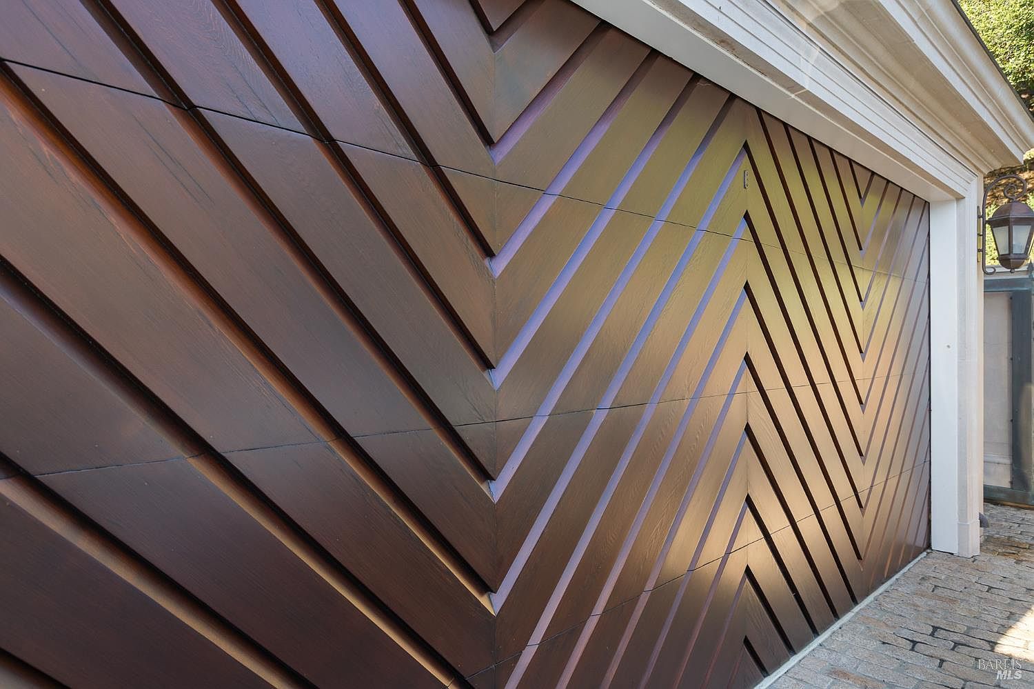 This image captures a close-up, angled perspective of a custom garage door featuring a striking chevron pattern created by dark-stained wood planks. The geometric design adds a sophisticated, modern architectural detail to the home's exterior, complemented by a classic lantern-style wall sconce mounted on the adjacent white trim. The perspective emphasizes the texture and depth of the wood grain and the precise craftsmanship of the paneling.