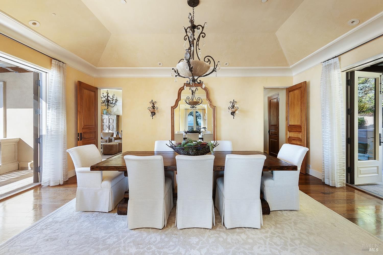 This elegant dining room features a large, rustic wooden table surrounded by six white slipcovered chairs, set atop a light-colored patterned area rug. The space is anchored by a grand, ornate iron chandelier hanging from a tray ceiling, with a large decorative mirror reflecting the room's warm, yellow-toned walls. Two sets of glass doors on either side provide natural light and access to outdoor spaces, creating a bright and sophisticated atmosphere.