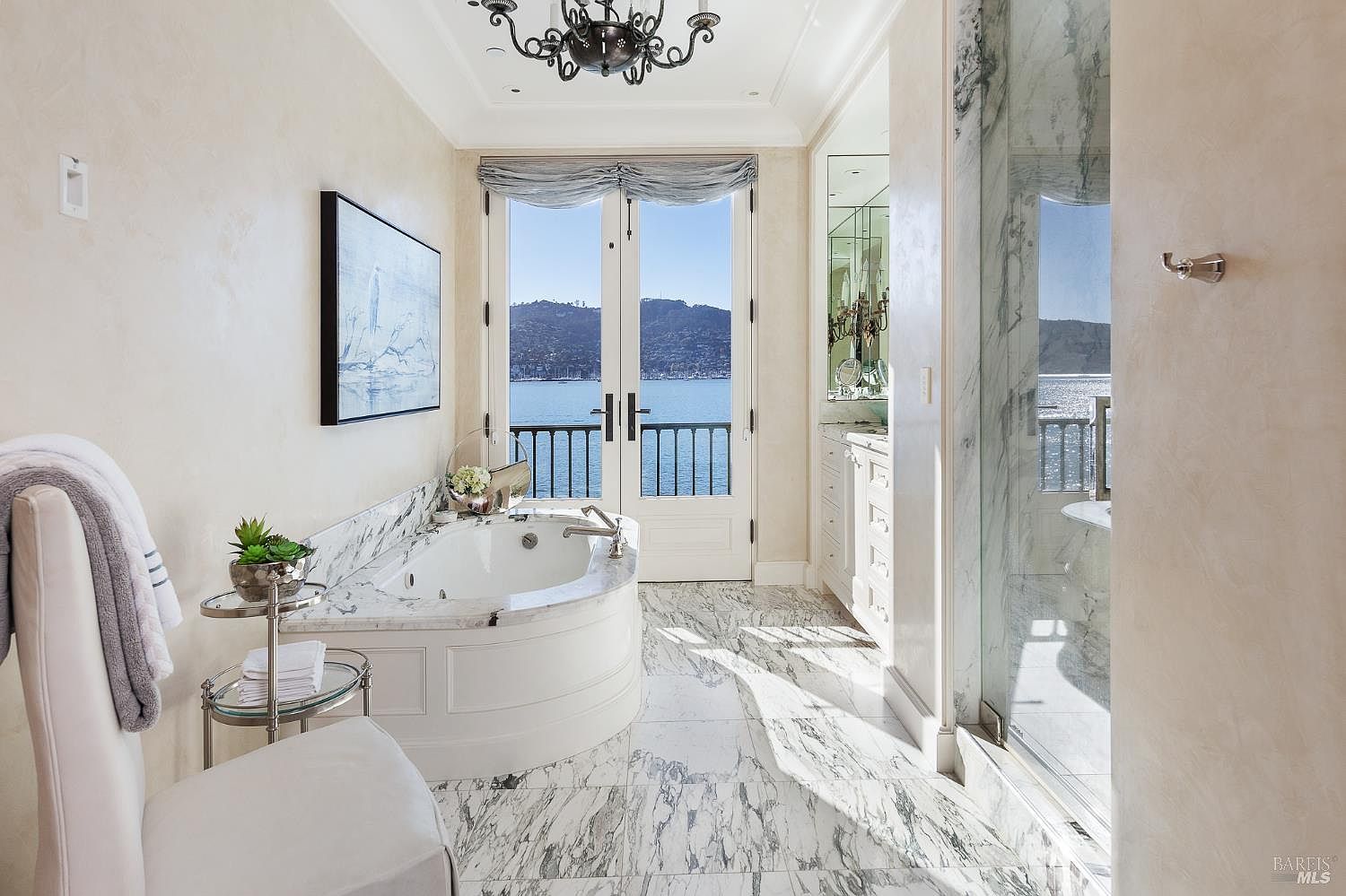 This luxurious primary bathroom features a deep soaking tub set within a stone-topped surround, positioned directly in front of French doors that offer expansive water views. The space is finished with patterned stone flooring and walls, a glass-enclosed shower, and a decorative chandelier, creating an elegant and serene atmosphere. The perspective is centered on the tub and the scenic outdoor vista, highlighting the high-end finishes and tranquil setting.