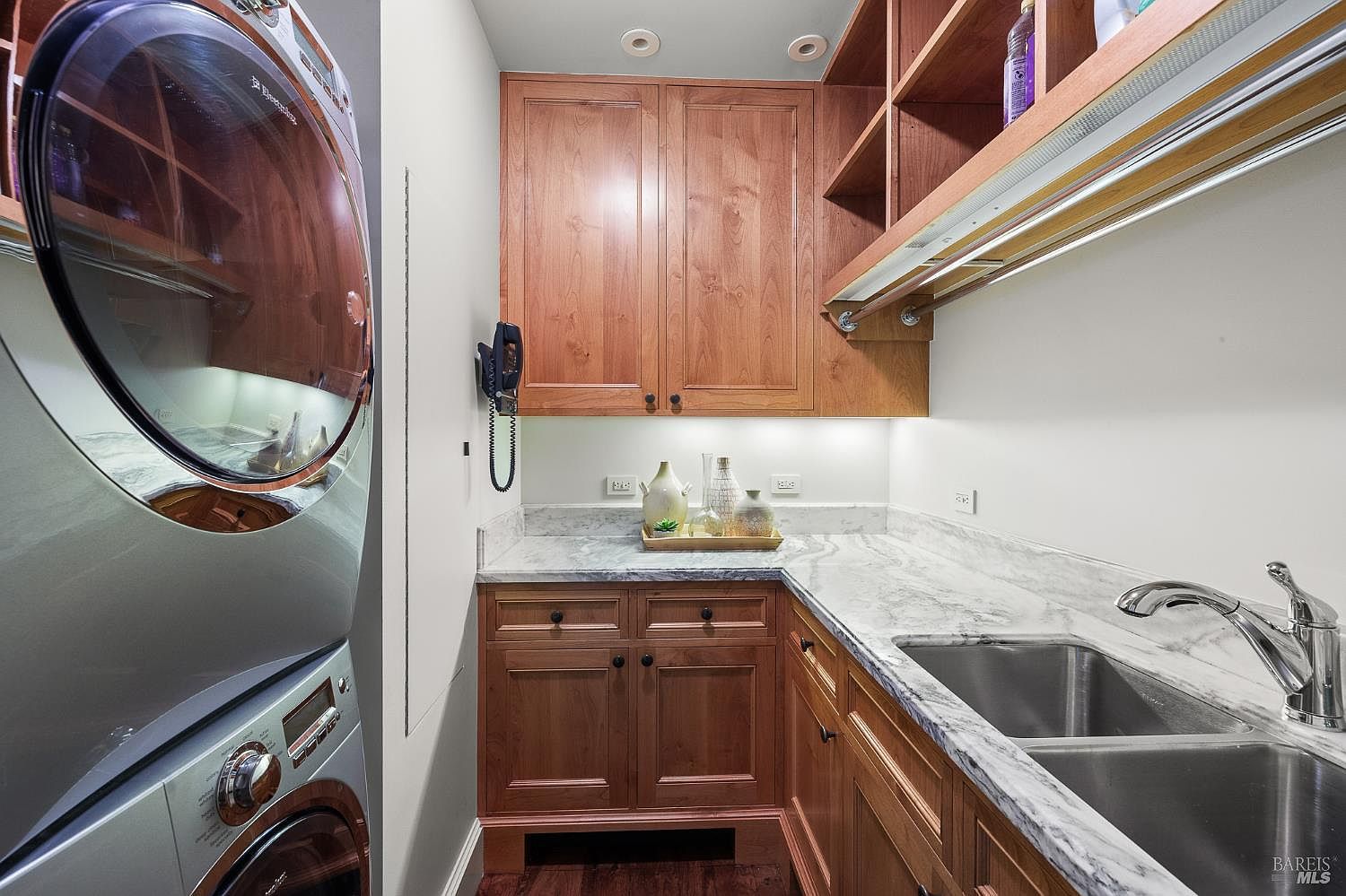 This functional laundry room features a stacked washer and dryer unit positioned next to a corner cabinet setup with a stone countertop and a double-basin sink. The space includes warm-toned wood cabinetry, open shelving, and under-cabinet lighting that illuminates the workspace. The perspective is a straight-on view, highlighting the efficient use of space and the clean, organized layout.