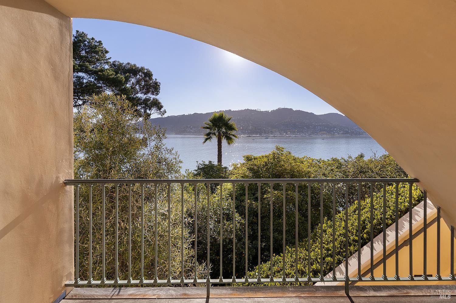 This balcony offers a serene, elevated view of a calm bay framed by a distinctive arched architectural opening. The space features a simple metal railing and a wooden deck, providing an unobstructed vantage point of the lush trees, a solitary palm, and the distant mountains across the water. The warm, neutral tones of the surrounding structure create a peaceful and inviting atmosphere.